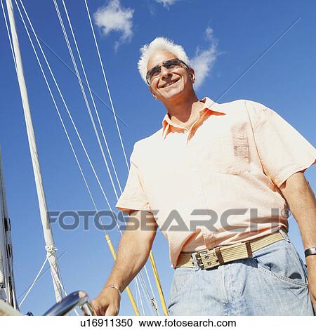 Stock Image - Low angle view of a mature man standing in a sailboat and smiling. Fotosearch