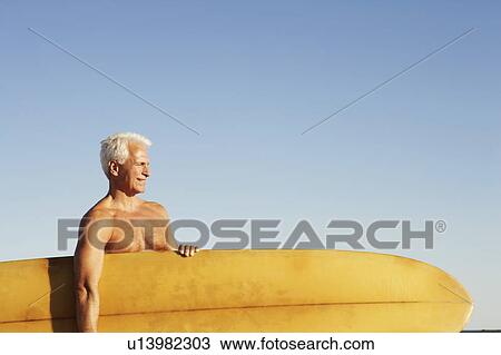 Stock Image - Mature man holding a surfboard under his arm. Fotosearch