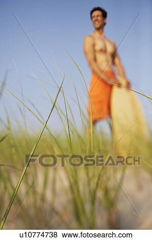 Stock Photo - Mature man standing on the beach and holding a wooden surfboard. Fotosearch