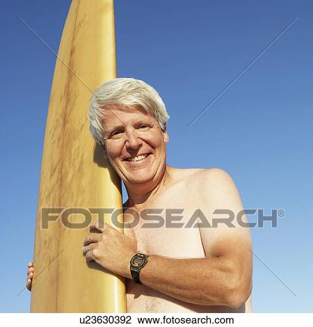 Stock Image - Portrait of a mature man holding a surfboard and smiling. Fotosearch