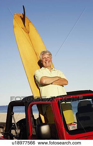 Stock Photograph - Portrait of a mature man leaning against a surfboard in a sports utility vehicle. Fotosearch