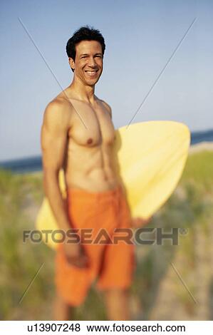 Stock Photo - Portrait of a mature man standing on the beach and holding a wooden surfboard. Fotosearch