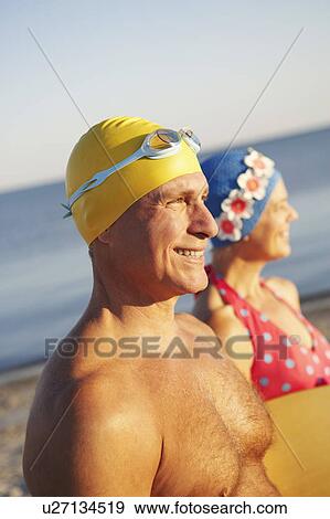 Stock Photo - Side profile of a mature man with a mature woman holding a surfboard on the beach. Fotosearch