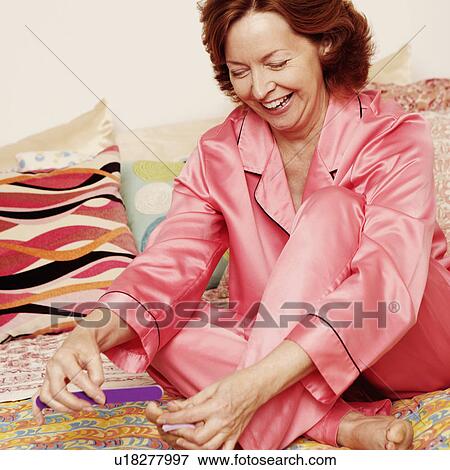 Stock Photo - Mature woman sitting on the bed and filing her toenails with a nail file. Fotosearch