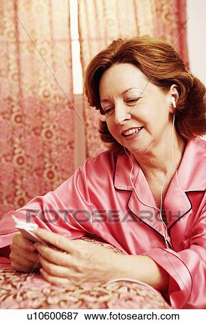Stock Photo - Mature woman leaning against a bed and listening to an MP3 player. Fotosearch