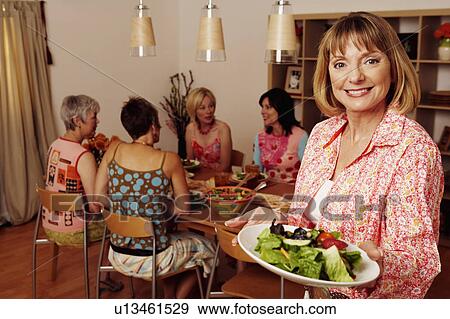 Stock Photo - Portrait of a mature woman holding a plate of salad and her friends sitting at a dining table in the background. Fotosearch