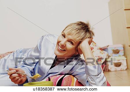Stock Photo - Portrait of a mature woman lying on the floor and eating corn flakes. Fotosearch