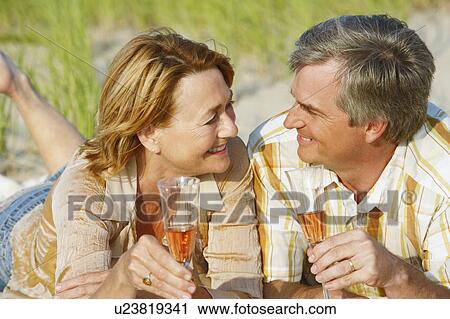 Stock Image - Close-up of a mature couple lying on the beach and holding champagne flutes. Fotosearch