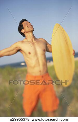 Stock Photo - Mature man standing on the beach and holding a wooden surfboard. Fotosearch