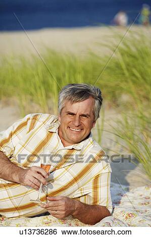 Stock Photograph - Portrait of a mature man lying on the beach and holding a champagne flute. Fotosearch