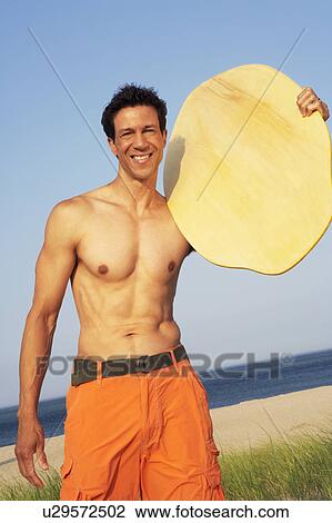 Stock Image - Portrait of a mature man standing on the beach and holding a wooden surfboard. Fotosearch