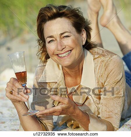 Stock Photography - Portrait of a mature woman lying on the beach and holding a champagne flute. Fotosearch
