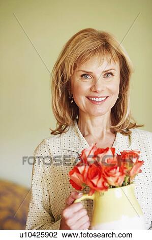 Stock Image - Portrait of a mature woman holding a pitcher with flowers and smiling. Fotosearch