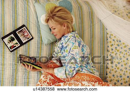 Stock Photo - High angle view of a mature woman lying in bed and looking at a photo album. Fotosearch