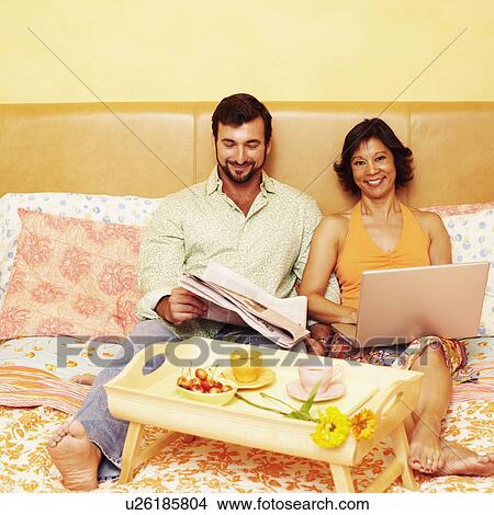 Mature man reading a newspaper with a mature woman sitting with a laptop on the bed View Large Photo Image Picture - Mature man reading a newspaper with a mature woman sitting with a laptop on the bed. Fotosearch