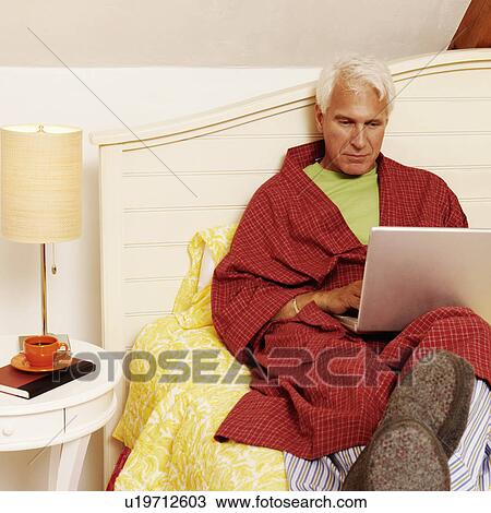 Stock Image - Mature man sitting on the bed and using a laptop. Fotosearch