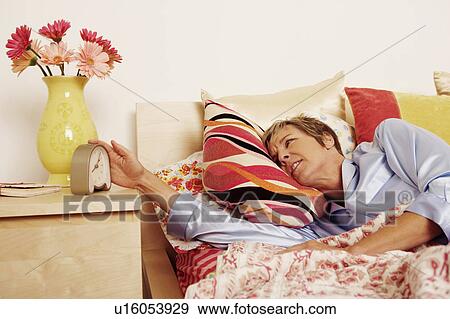 Mature woman lying in bed and reaching for an alarm clock View Large Photo Image Stock Photo - Mature woman lying in bed and reaching for an alarm clock. Fotosearch