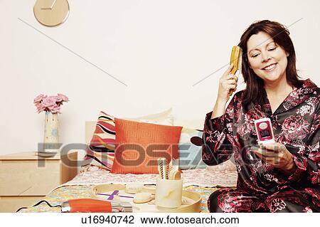 Stock Image - Mature woman sitting on the bed with a flip phone and combing her hair. Fotosearch