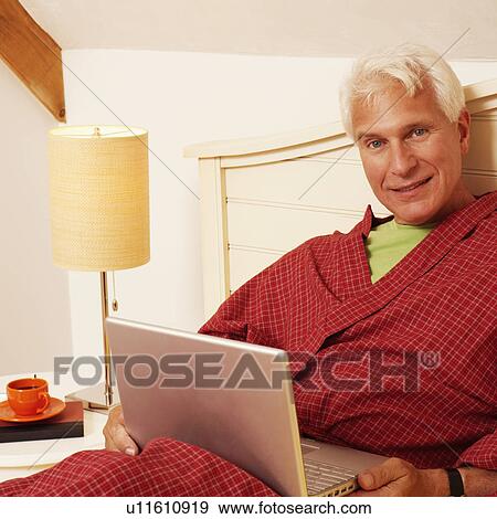 Stock Photo - Portrait of a mature man leaning on the bed with a laptop. Fotosearch