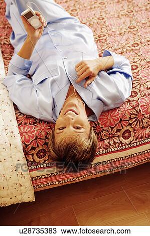 Stock Image - Portrait of a mature woman lying on the bed listening to an MP3 player. Fotosearch