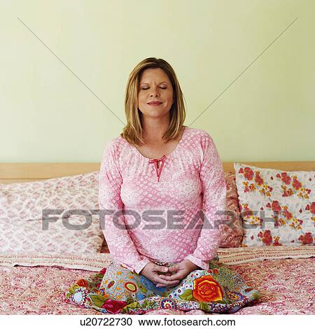 Stock Image - Mature woman sitting on the bed and meditating. Fotosearch