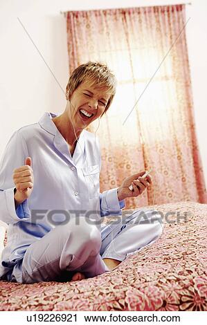 Stock Image - Mature woman sitting on the bed listening to an MP3 Player. Fotosearch