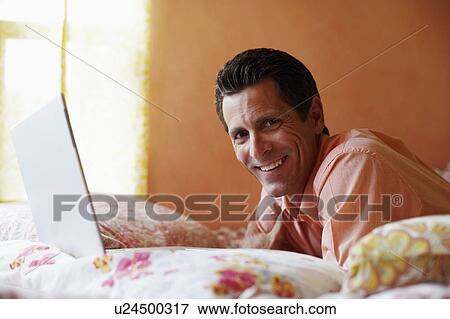 Stock Photo - Portrait of a mature man lying on the bed with a laptop and smiling. Fotosearch