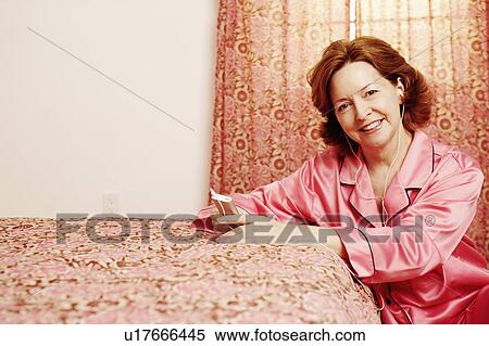 Stock Photography - Portrait of a mature woman leaning against the bed and listening to an MP3 player. Fotosearch