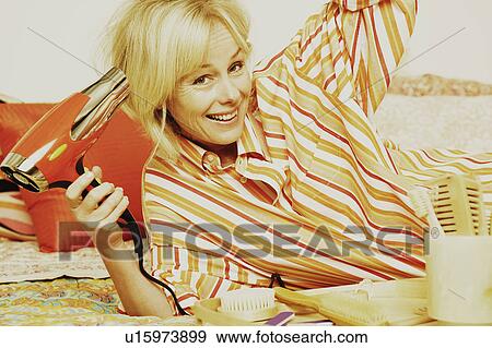 Stock Photo - Portrait of a mature woman lying in bed and drying her hair with a hair dryer. Fotosearch