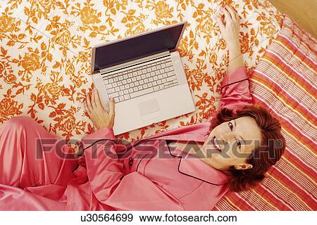 Portrait of a mature woman lying in bed with a laptop and smiling View Large Photo Image Stock Photo - Portrait of a mature woman lying in bed with a laptop and smiling. Fotosearch