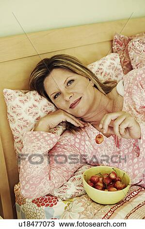 Stock Image - Portrait of a mature woman lying on the bed with a bowl of cherries. Fotosearch
