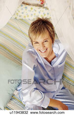 Stock Image - Portrait of a mature woman sitting on the bed and smiling. Fotosearch