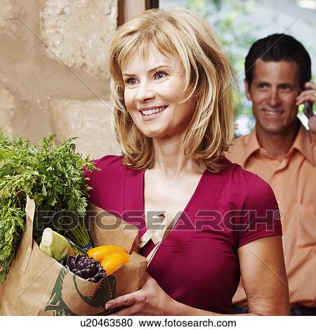 Close-up of a mature woman holding a shopping bag with a mature man talking on a mobile phone behind him View Large Photo Image Stock Image - Close-up of a mature woman holding a shopping bag with a mature man talking on a mobile phone behind him. Fotosearch
