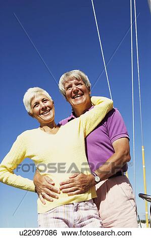 Stock Photograph - Low angle view of a mature man embracing a mature woman from behind in a boat and smiling. Fotosearch