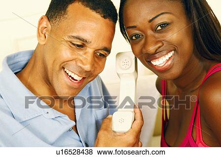 Stock Photo - Close-up of a mature man and a mid adult woman listening to a telephone receiver and smiling. Fotosearch