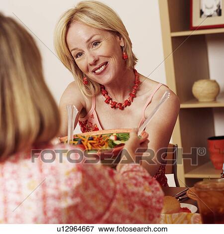 Stock Photo - Mature woman giving a bowl of salad to her friend and smiling. Fotosearch