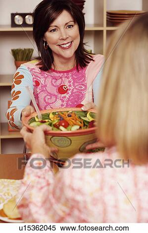 Stock Photography - Mature woman giving a bowl of salad to her friend and smiling. Fotosearch