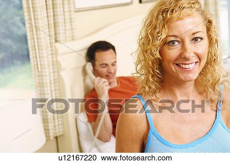 Stock Image - Close-up of a mature woman smiling with a mature man talking on the telephone in the background. Fotosearch