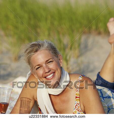 Picture - Portrait of a mature woman lying on the beach and holding a champagne flute. Fotosearch