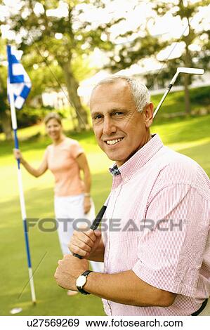 Portrait of a mature man holding a golf club and a mature woman holding a golf flag in the background View Large Photo Image Stock Photo - Portrait of a mature man holding a golf club and a mature woman holding a golf flag in the background. Fotosearch