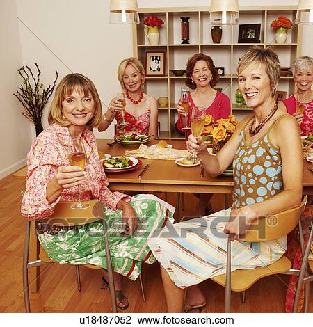 Stock Image - Portrait of a group of mature adult woman sitting at dining table holding champagne flutes. Fotosearch