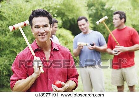 Stock Image - Portrait of a mature man holding a croquet mallet and a ball with his friends in the background. Fotosearch