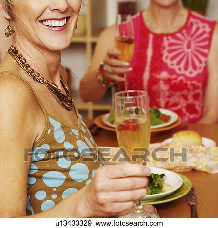 Stock Photo - Close-up of a mature woman sitting at the dining table with her friend and holding a champagne flute. Fotosearch