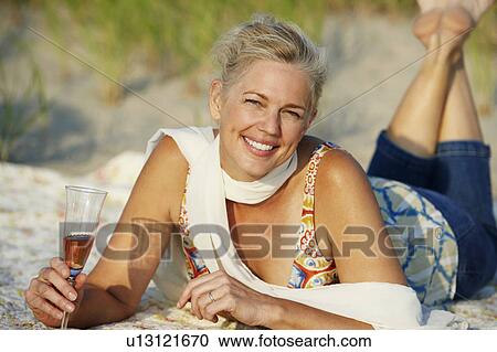 Stock Image - Portrait of a mature woman lying on the beach and holding a champagne flute. Fotosearch