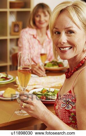 Stock Image - Portrait of a mature woman sitting at the dining table with her friend and holding a champagne flute. Fotosearch