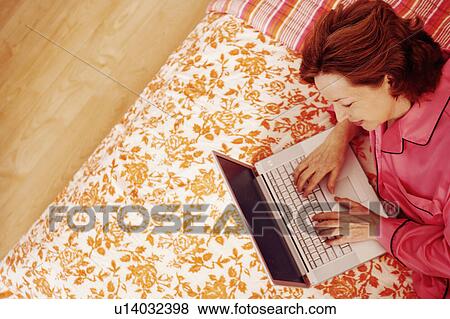 Stock Photo - High angle view of a mature woman lying in bed using a laptop. Fotosearch