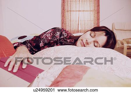 Stock Image - Mature woman sleeping on the bed with her hand on an open book. Fotosearch