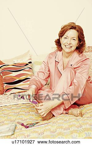 Stock Photo - Portrait of a mature woman sitting on the bed and filing her toenails with a nail file. Fotosearch