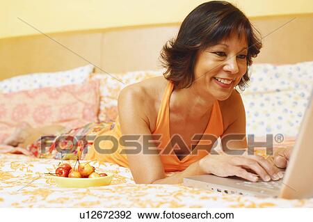 Stock Image - Close-up of a mature woman lying on the bed and using a laptop. Fotosearch