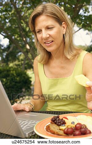 Picture - Close-up of a mature woman using a laptop and eating fruit salad. Fotosearch